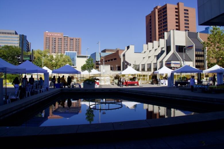 Several booths are set up outdoors in a city space with a water feature. Tall buildings are visible behind them.