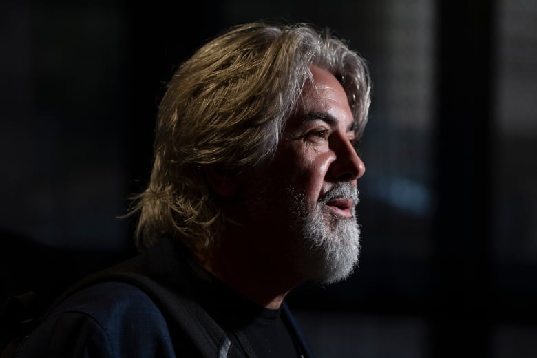 A man with long silver hair and a beard speaks to reporters who are not pictured here.