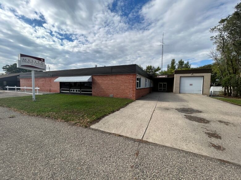 A brick building with a striped awning over a window and a cement pad driveway is shown. There is a white garage door entrance on screen right and a sign which says Melita Health Centre.