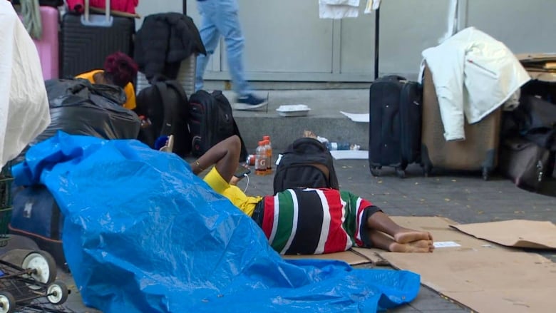 A person can be seen sleeping on the ground, with cardboard underneath them. Luggage can be seen nearby.