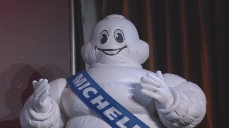 A white puffy mascot with a blue sash saying 