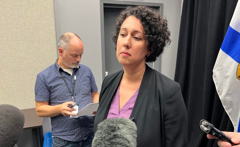 A woman with curly hair and wearing a blazer stands in front of reporters holding microphones.