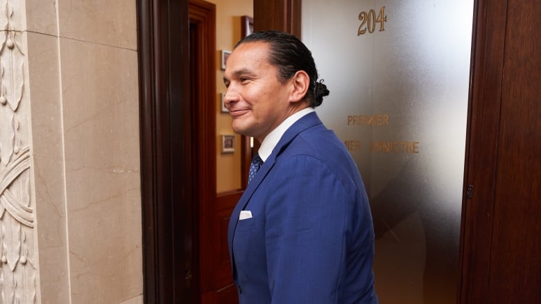 A politician in a ponytail and blue suit enters a room with the word 'premier' on the door.