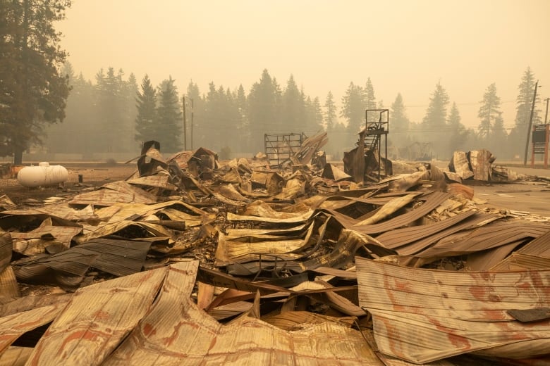 Flattened and warped metal from a burnt building is shown beneath a sky that is yellow from wildfire smoke.