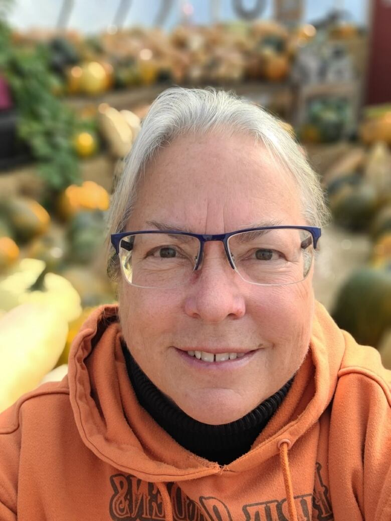 a woman with grey hair in an orange hoodie stands stands in front of a background of pumpkins and smiles