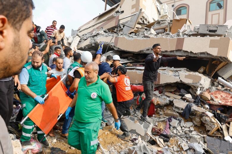 Two men wearing green uniforms carry an orange canvas stretcher in front of a crowd of people on top of a pile of concrete rubble and a collapsed building. 