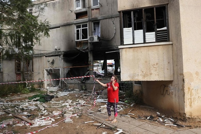 A woman in a red t-shirt stands outside a heavily damaged building speaking on a mobile phone.