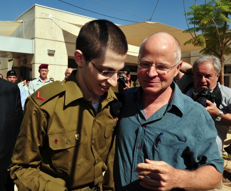 A young man in a green military uniform and wearing glasses looks downward with a smile on his face as another older man, in a blue shirt and glasses, walks beside him with an arm wrapped around his back. 