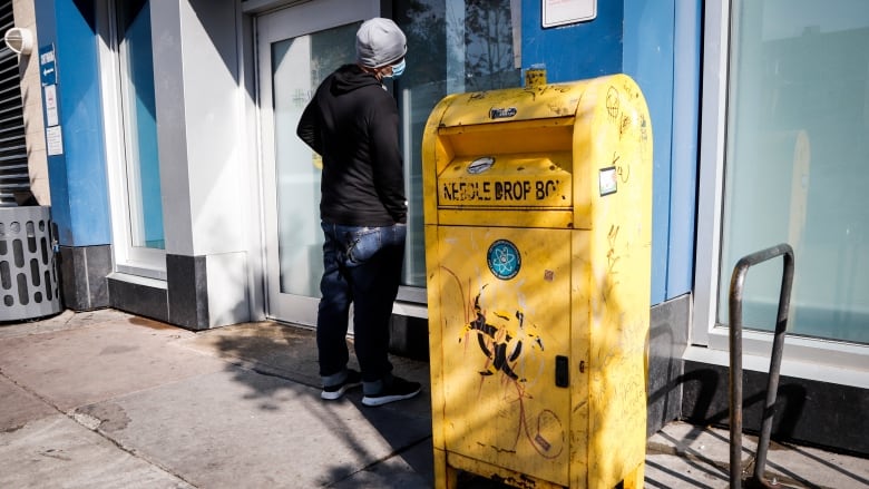 A man wearing a toque and a mask stands outside a building where there is a bin outside that reads 