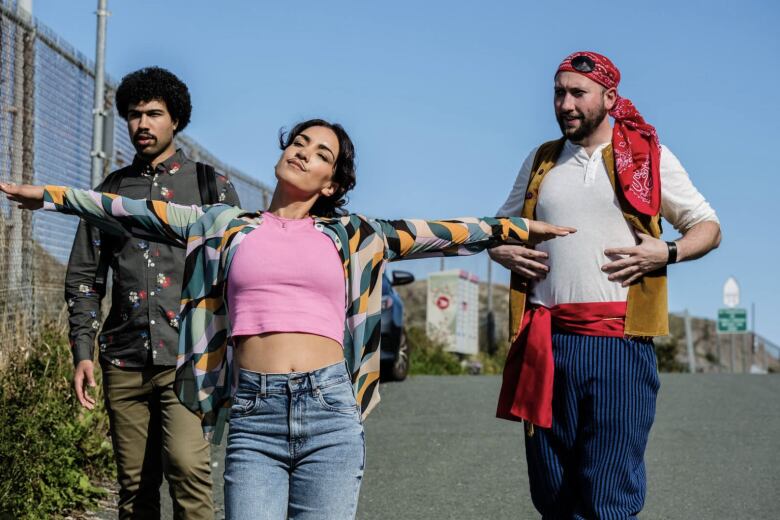 A woman with black hair, pink t-shirt, blue jeans and a multi-coloured shirt has her arms extended walking in front of two men, one with black hair on the left in olive pants, a green shirt with a pattern and on the right is a man with a red kerchief, white shirt, gold vest, blue pants and a red sash around his waist.