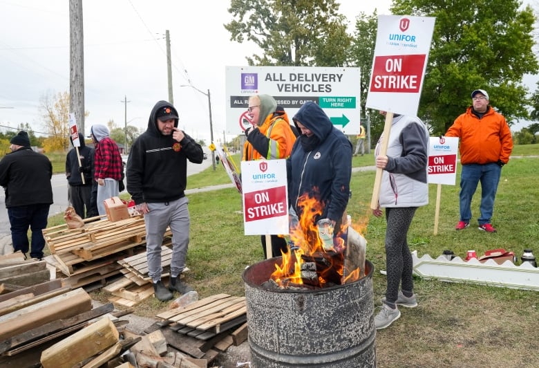 A fire in a barrel, surrounded by people holding picketing signs that say 'On Strike'.