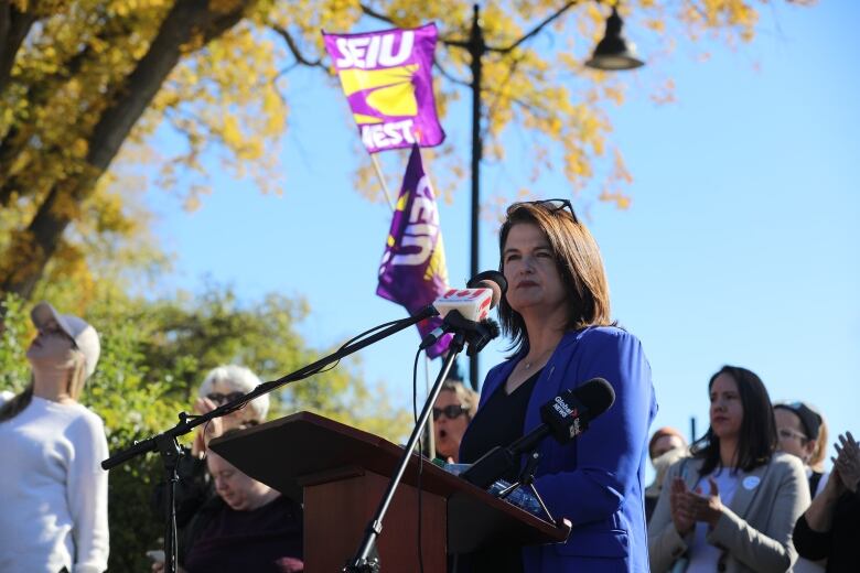 A woman in a blue blazer and a black shirt stands at a podium outside the provincial legislature. 