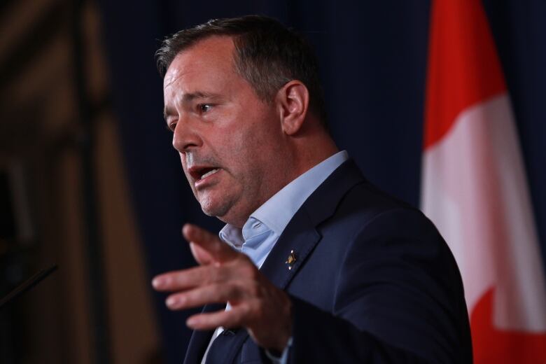 A man in a suit gestures while standing in front of a Canadian flag.