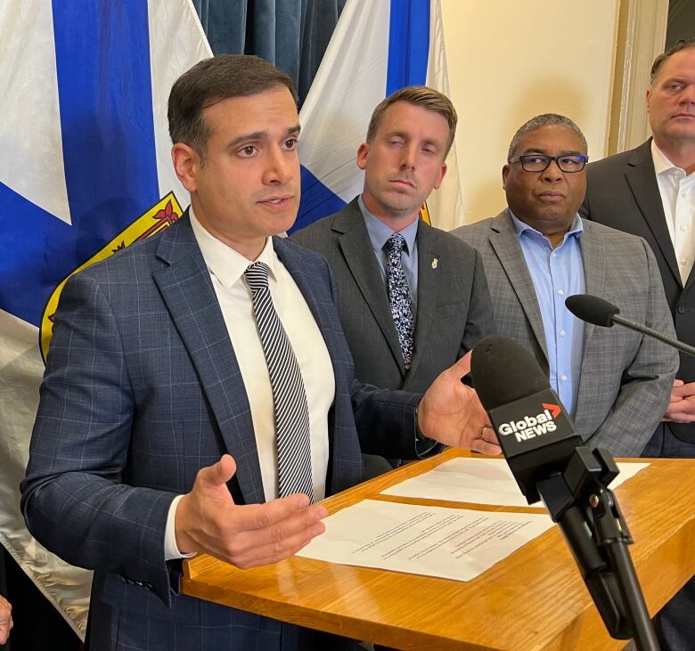 A man in a suit stands behind a microphone, in front of a Nova Scotia flag. Three other men wearing suits are standing to his left. 