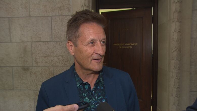 A man in a blue blazer and floral print shirt speaks to members of the media.