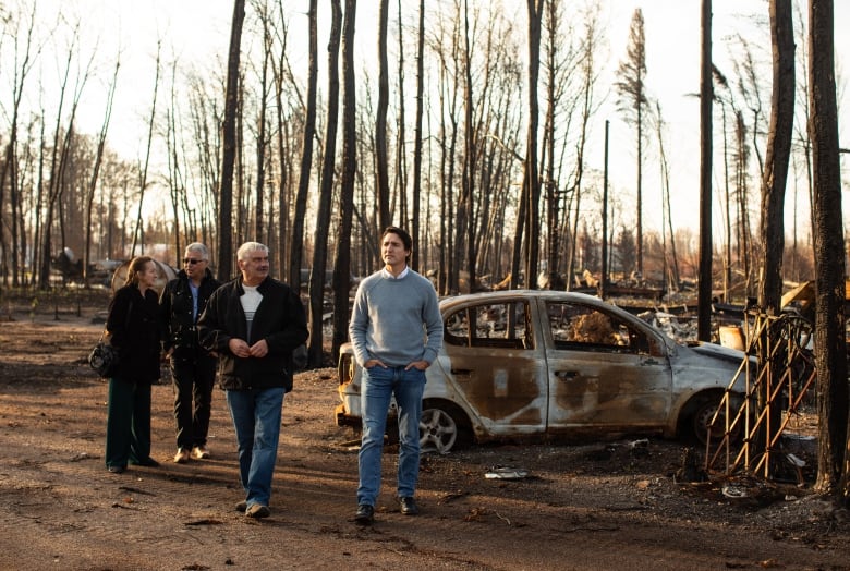 four people stand in front of a burned vehicle in front of many burnt trees