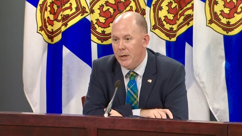 A man wearing a suit sits behind a long wooden podium in front of a screen and Nova Scotia flags.