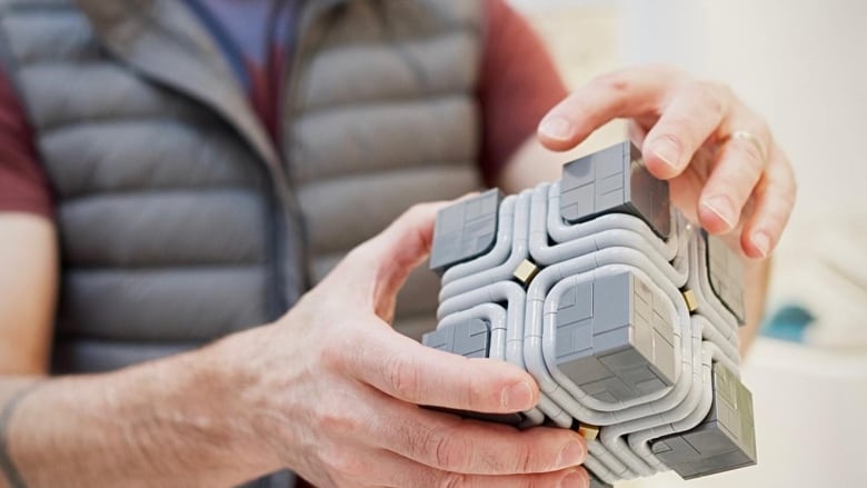 A man holds a small gray cube made from Lego blocks.