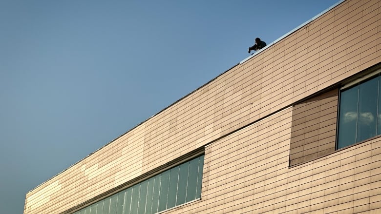 A construction worker attaches edging material high up on the rooftop of a gold coloured building.