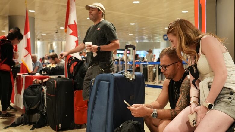 A man and woman checking their phones sit by their suitcases in a line at an airport filled with Canadian flags as other people wait with their luggage. 