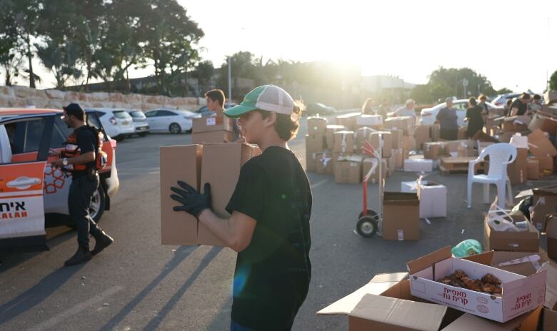 A person wearing a dark t-shirt, a ball gap and dark gloves, carries a box while surrounded by other boxes in a parking lot. 