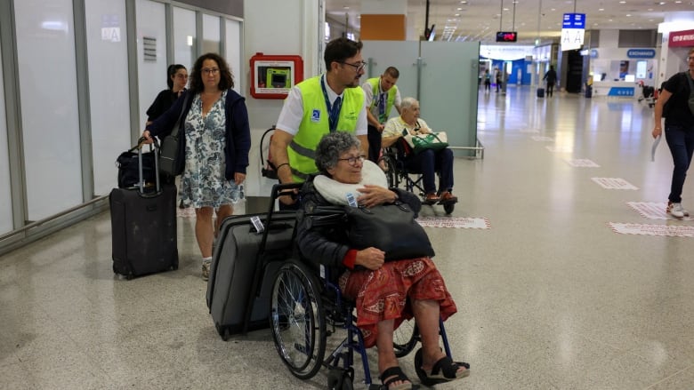 A man in a yellow vest rolls a woman in a wheel chair and her suit case into an airport terminal.