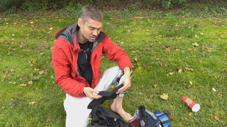 A man in a red jacket is shown kneeling down in bare feet, showing his socks both in his hands with other pairs on the grass beside him.