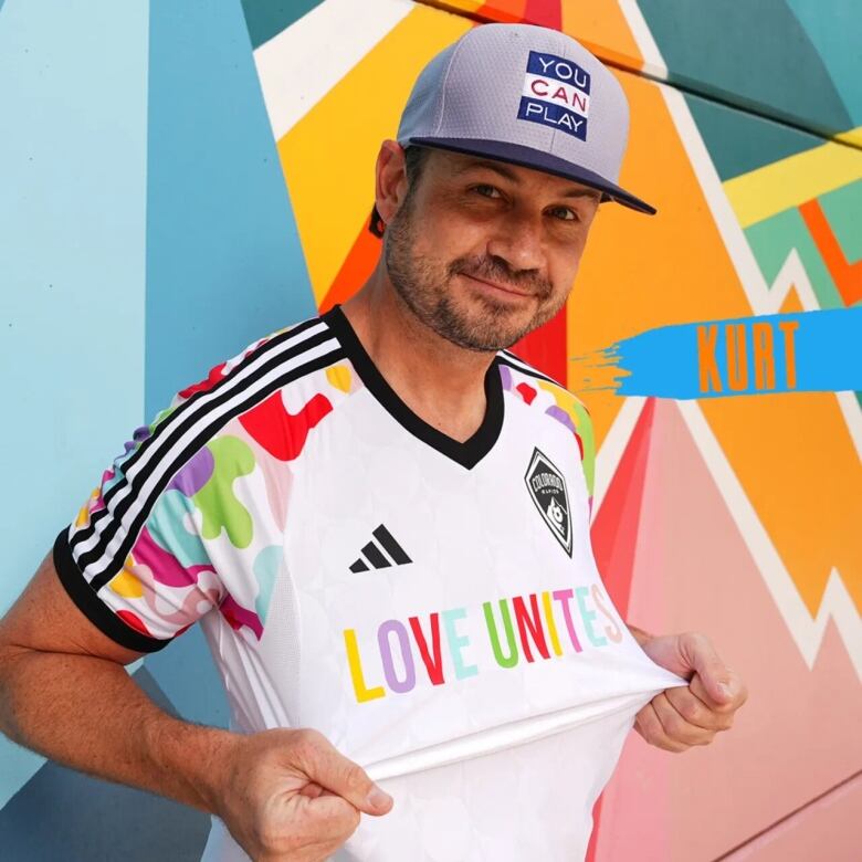 A man in a baseball cap holds out the front of his T-shirt, reads Love Unites in multi-coloured letters.