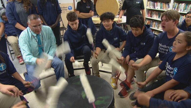 a adult wearing a light dress shirt with tie, is sitting with young students around a drum and singing.