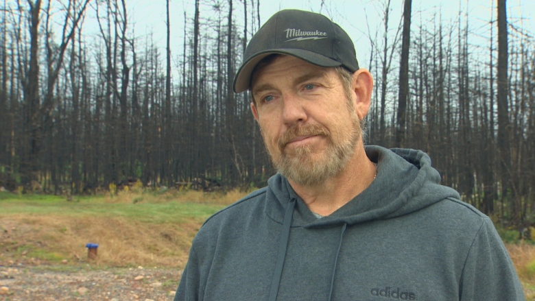 A man with a beard wears a grey sweatshirt and ball cap. Behind him is a vacant lot and burnt trees. 