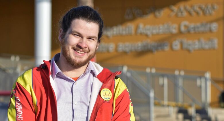 A man with long hair that is tied back and a purple dress shirt and yellow and red jacket smiles at the camera in a photo taken outside. 