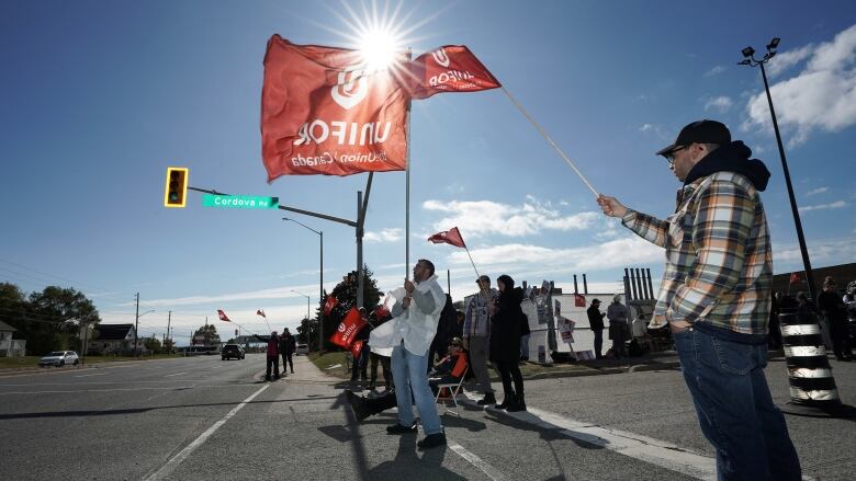 Workers wave Unifor flags on a picket line on a sunny fall day.