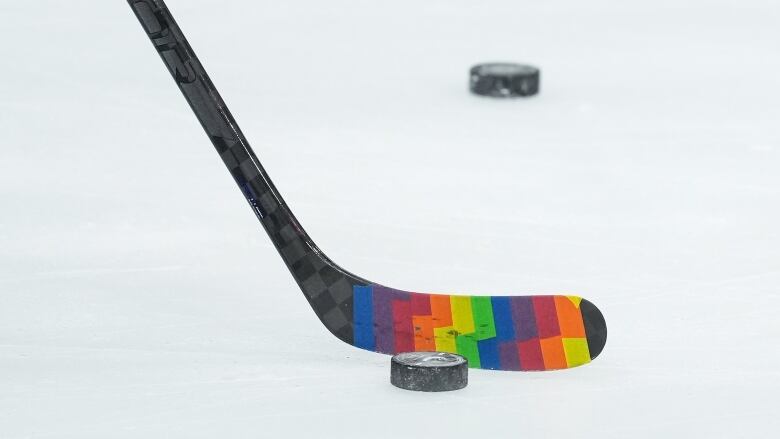 Close-up of a hockey stick wrapped in rainbow-coloured Pride Tape approaching a puck.