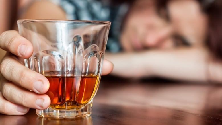 Glass of alcohol on a table in the foreground, held by a woman with her head on the table who is out of focus in the background