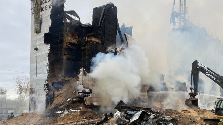 Smoke rises from a tall, scorched building. Scrap metal pieces sit in front of the building while grain spills from the building's centre.