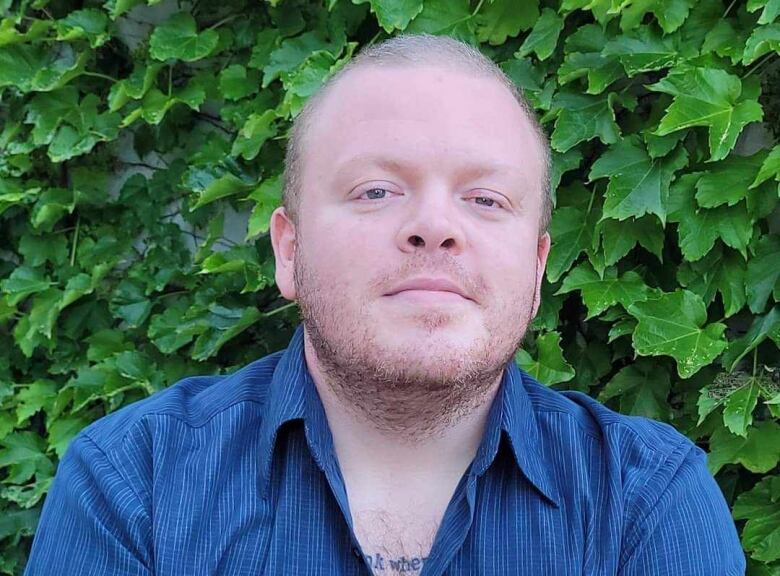 A man wears a blue button up blouse standing in front of green leaves.