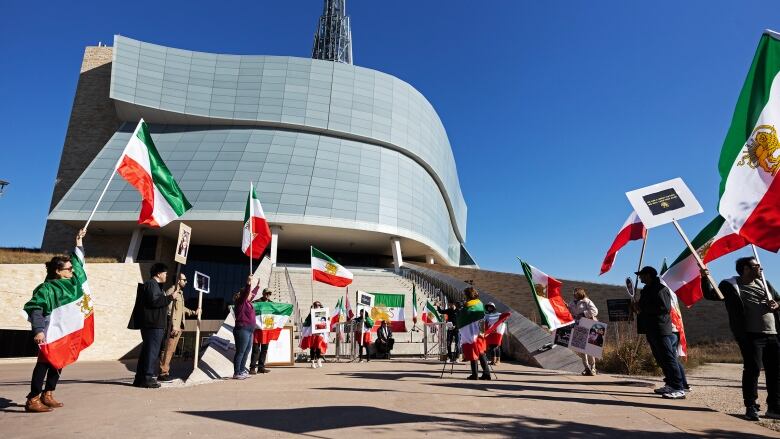 About 10 people are seen standing outside the Canadian Museum for Human Rights, waving flags and holding up signs.