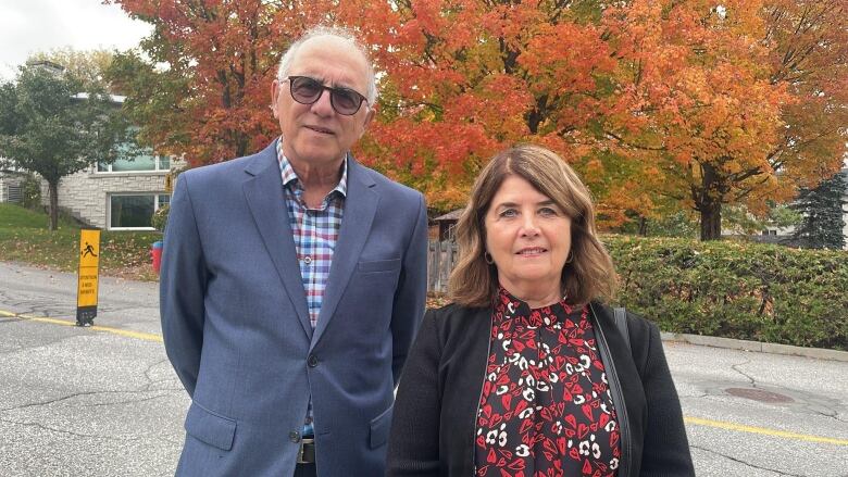 Man and woman standing and smiling in front of a street, with colourful tree behind them.