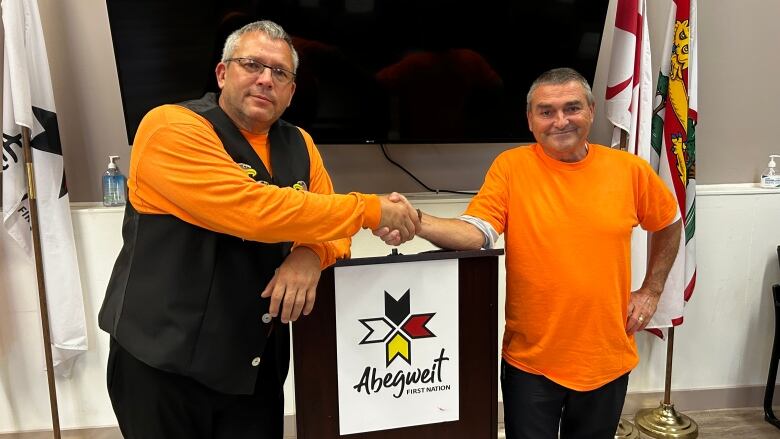 Two men wearing orange pose for a picture shaking hands in front of a podium with a sign that says