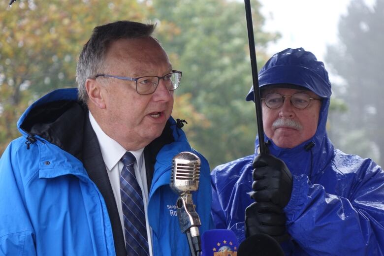 A man wearing glasses speaks into a microphone under an umbrella.