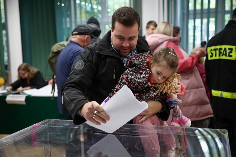 An adult holds a child as they put papers into a clear box.