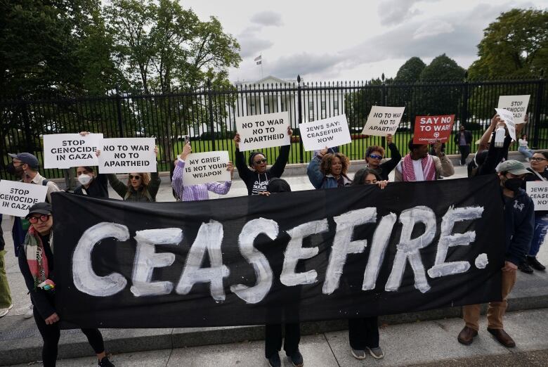 Several people hold signs in front of the fence outside the White House grounds in Washington, while others extend a banner that reads 'Ceasefire.'