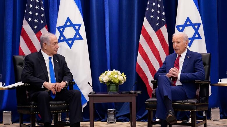 Two men in suits are seen sitting in chairs turned to each other, with American and Israel flags and drapes in the background.