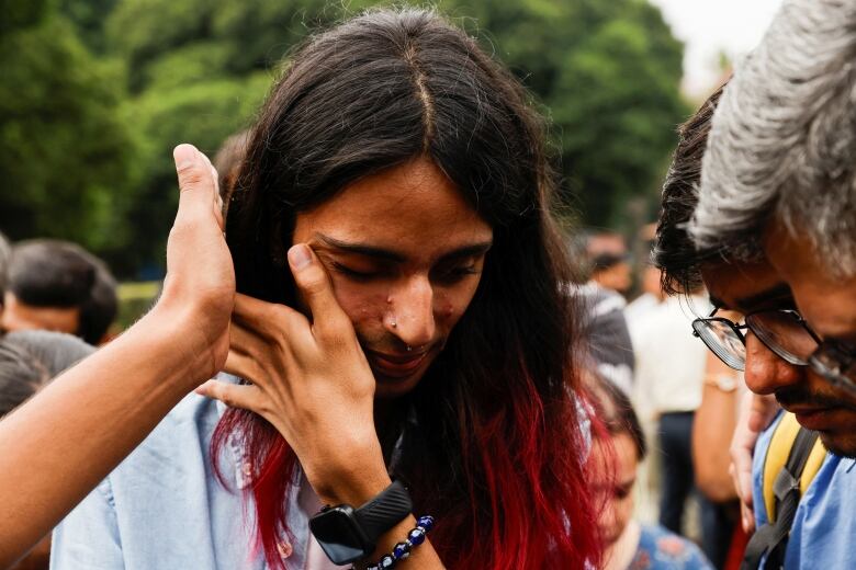 A person with pink shoulder-length hair wipes a tear from their eye.