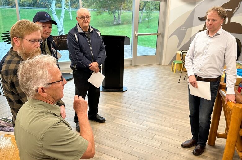 A man with a white shirt stands on the right while on the left, three people listen to a fourth man who is gesturing with his hand and speaking.