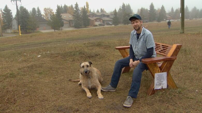 A man sits on a bench in a field with a dog  lying in front of him.