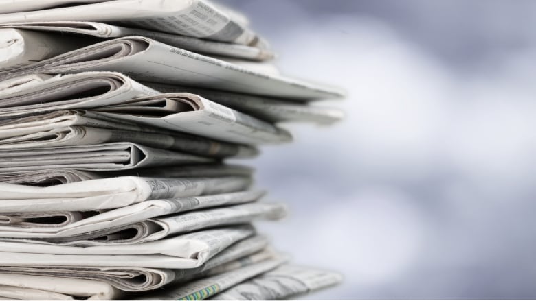 A pile of newspapers is stacked to the left side of the image, against a blurred background.