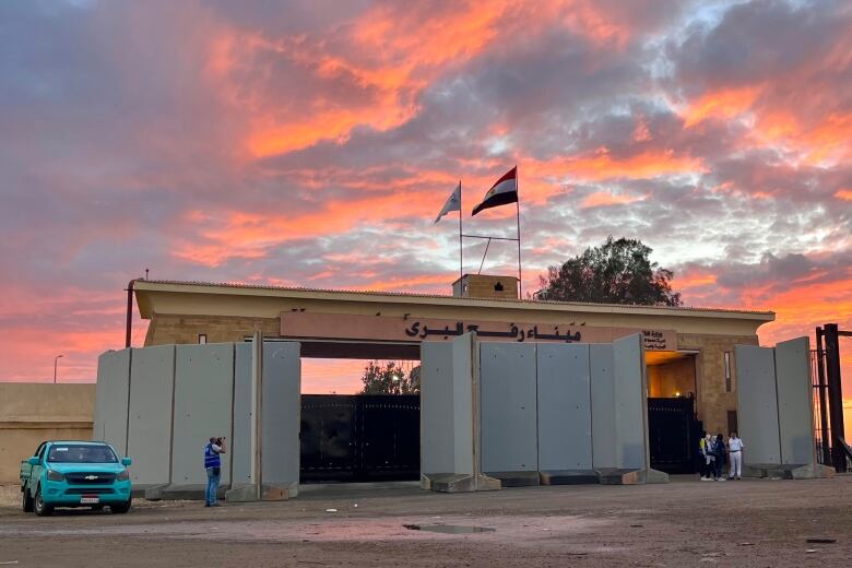 A man stands in front of a large, gated structure, with two flags on top, beneath a pink and blue, cloudy sky. 