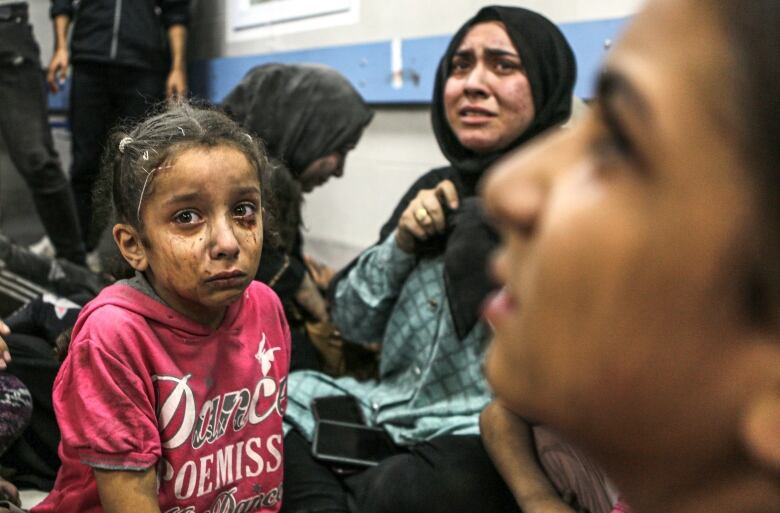 A girl, her face covered in mud and tears, sits in a corridor with a woman, who is also crying.