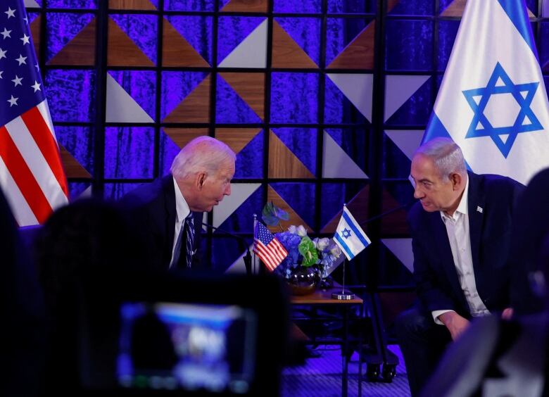 Two men in suits sitting in chairs tiled toward each other lean in for conversation. American and Israel flags are seen in the background.